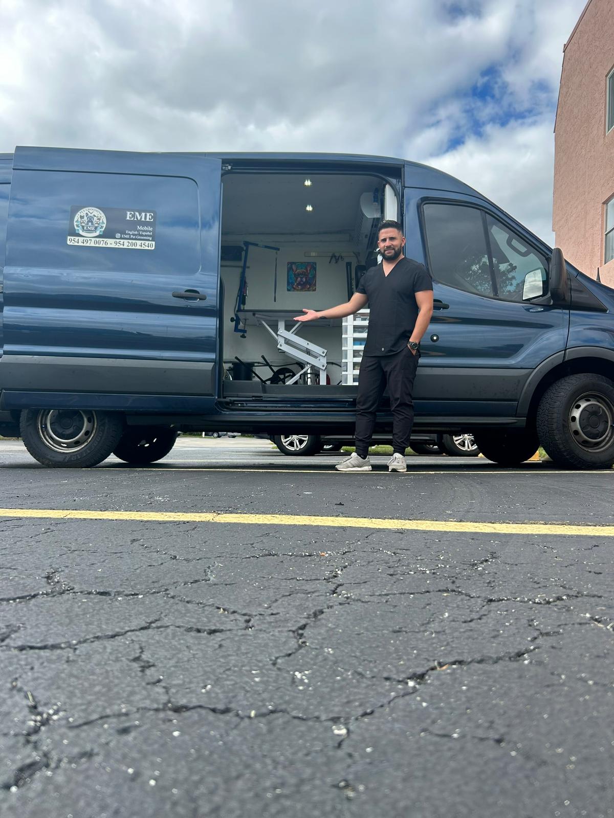 An image of a man standing in front of a van, the rear door of which is open, displaying a white massage table. He is wear...